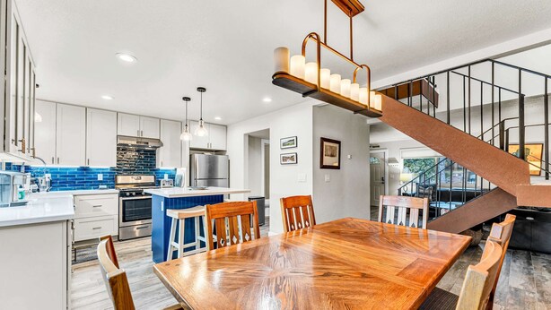 A modern kitchen with white cabinets, a blue backsplash, and stainless steel appliances. There is a wooden dining table with chairs, a staircase with a wooden railing, and pendant lights above the table. - Lake Tahoe Getaway – Close to Beach & Ski Slopes! (Tahoe City)