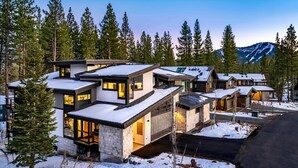 Un edificio residencial moderno de varios pisos, con grandes ventanales y un techo cubierto de nieve, rodeado de altos pinos y una montaña al fondo.