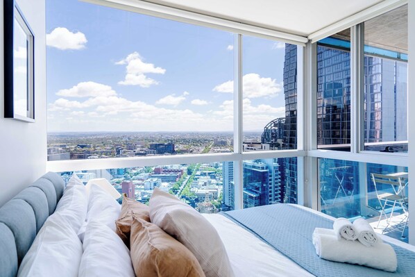 A bedroom with a large window, a bed with pillows, and a view of a cityscape.