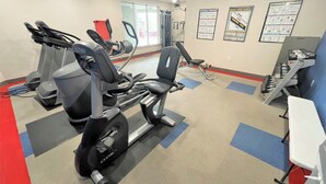 A gym with treadmills, a stationary bike, and a weight bench. There are posters on the wall and a white table with weights.
