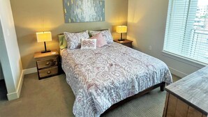 A bedroom with a large bed, two bedside tables with lamps, a nightstand, a wall-mounted artwork, and a window with blinds.