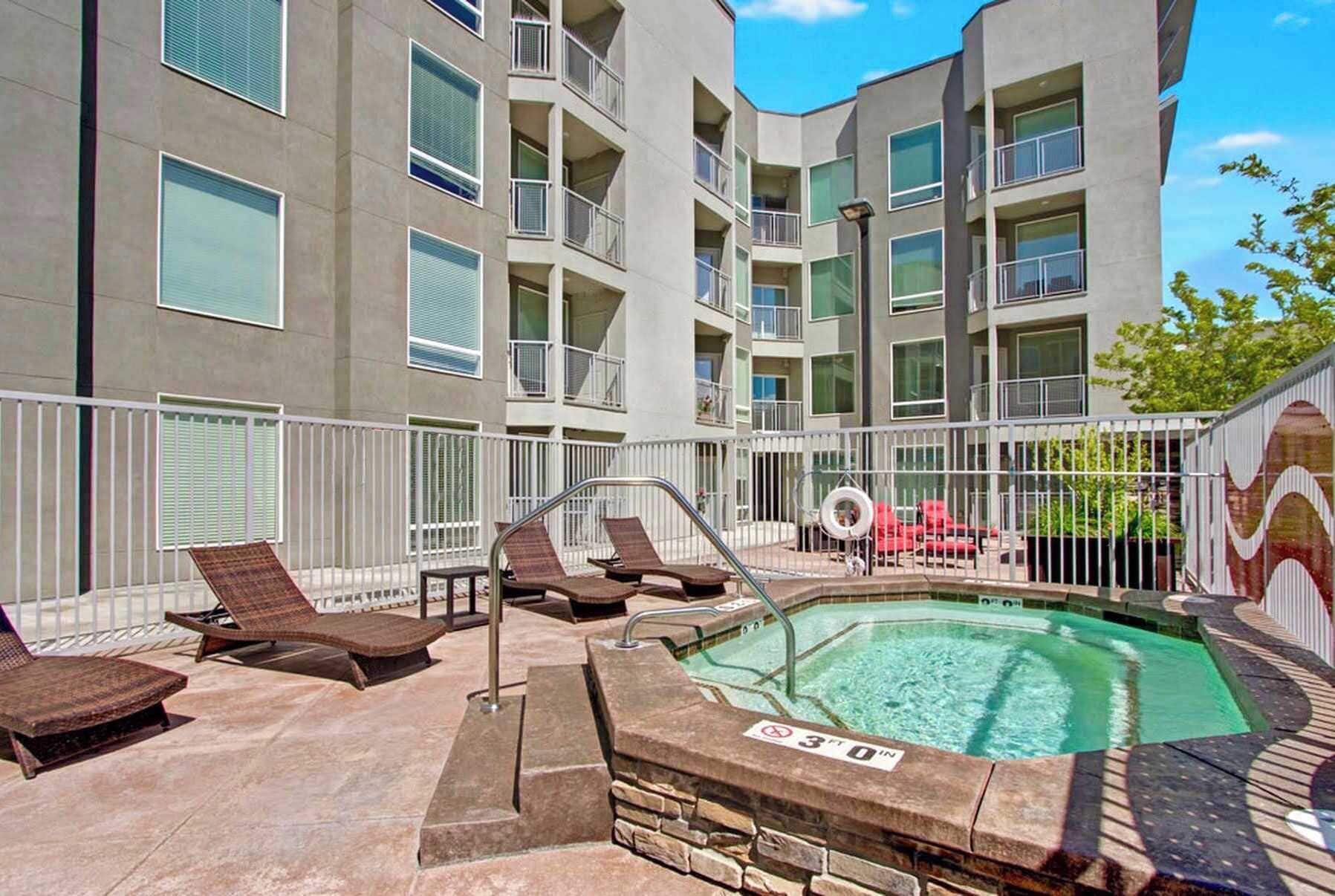 A pool area with lounge chairs and a hot tub, surrounded by a building with balconies.