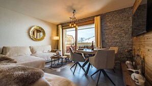 A cozy living room with a sofa, a dining table with chairs, a fireplace, and a large window with curtains.