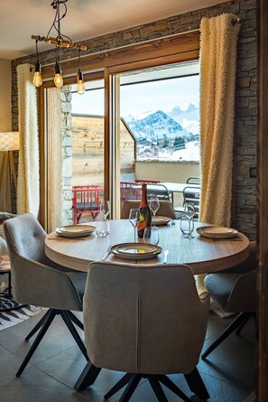 Un coin repas avec une table ronde dressée pour quatre personnes, une bouteille de vin et une vue sur les montagnes par de grandes fenêtres.