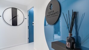 Un vestíbul d'hotel modern amb un disseny minimalista, que presenta un mirall rodó, un rellotge digital, un cartell de 'Sofia Apartments Italian Holidays Experiences' i una ampolla de perfum decorativa.