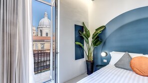 A bedroom with a bed, a potted plant, a wall-mounted light, and a view of a building through a window. - CORSO UMBERTO 228 (Napoli)