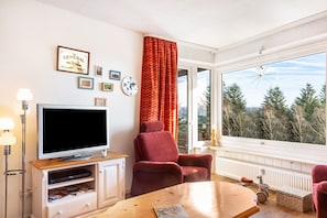 Una sala de estar con televisor, un sillón rojo, una mesa de centro de madera y un ventanal con vistas a árboles.