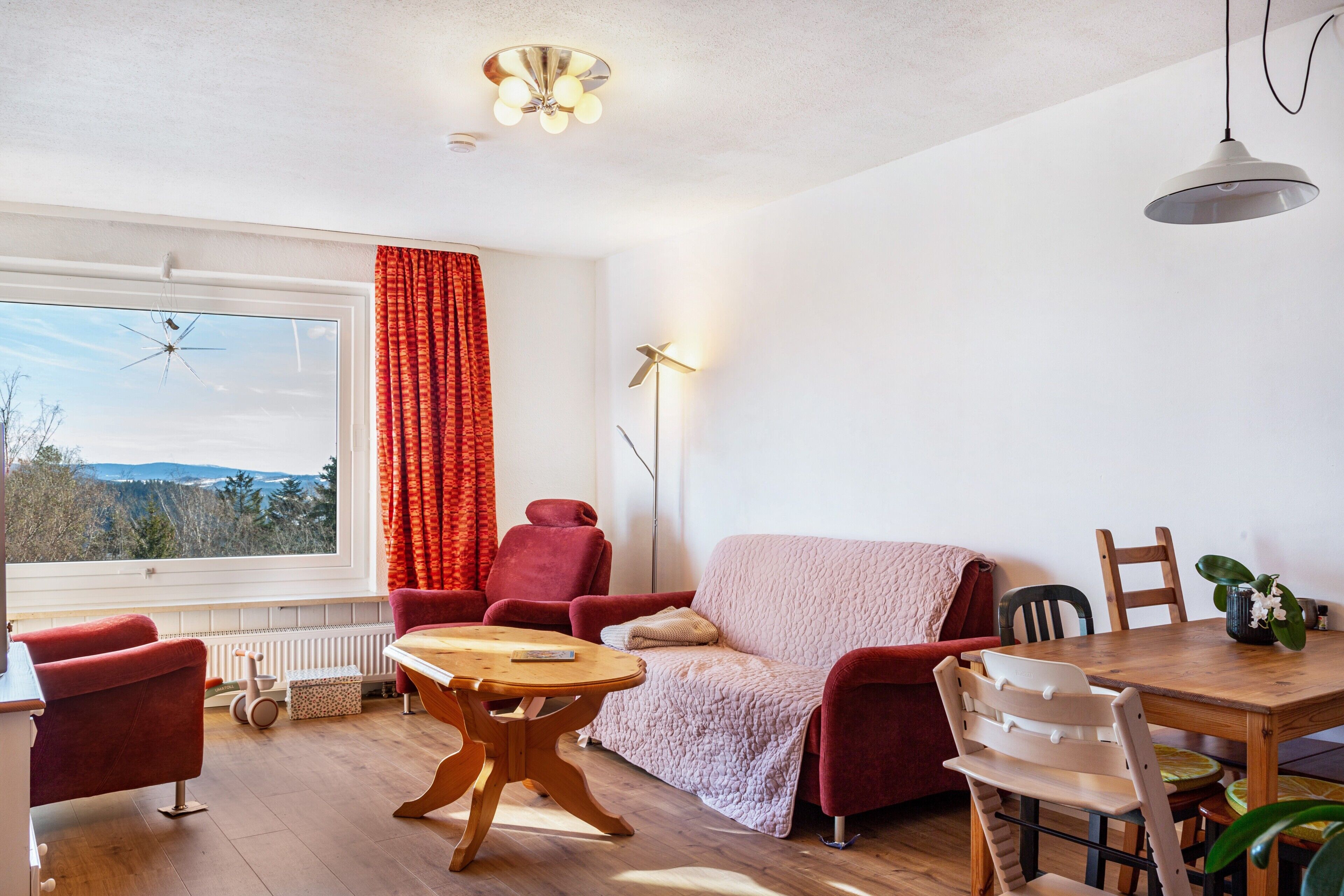 Un salon doté d’une grande fenêtre, de rideaux rouges, d’un canapé rouge, d’une table basse en bois et d’une table à manger avec des chaises.