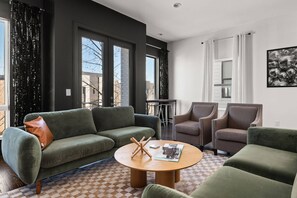 Una sala de estar moderna con un sofá verde, dos sillones, una mesa de centro y ventanas grandes con cortinas negras.
