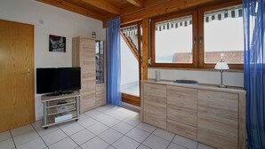 Een kamer met een houten plafond, een televisie op een standaard, een kast, een raam met gordijnen, en een deur.