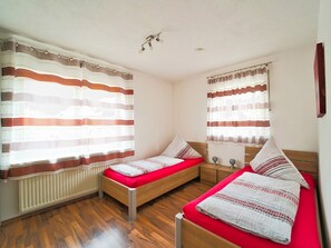 Habitación con dos camas, cada una con colchón rojo y ropa de cama blanca, flanqueada por ventanas con cortinas a rayas.