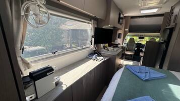 Un intérieur de camping-car compact comprenant un coin cuisine, un lit avec une couverture verte, une petite table avec un évier et une fenêtre avec des rideaux.