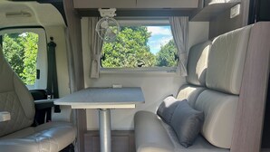 A compact RV interior with a dining table, cushioned seating, and a window offering a view of greenery.