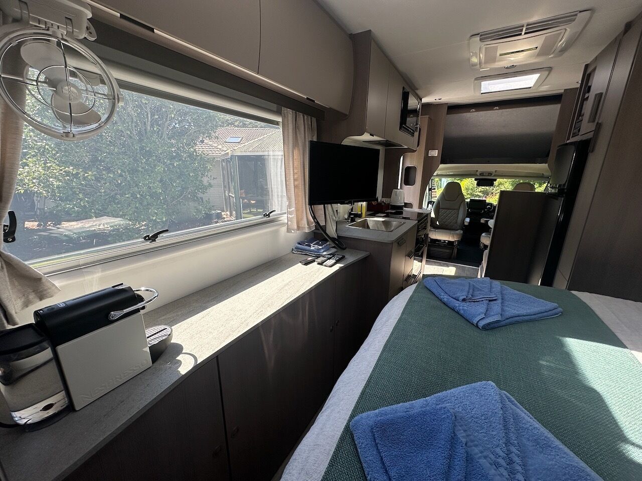 Interior compacto de RV com cozinha compacta, uma cama com toalhas azuis, uma TV e janela com vista para o verde.