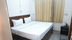 Una cama bien hecha con sábanas blancas y dos almohadas, cabecera de madera, mesita de noche con tetera y tazas, y cortinas beige en una ventana.