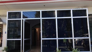 Entrada de un edificio con amplias puertas de vidrio, plantas en macetas y un coche reflejado.