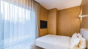 Habitación de hotel con un ventanal grande, una cama con ropa blanca, un televisor de pantalla plana en la pared y un cabecero de madera.