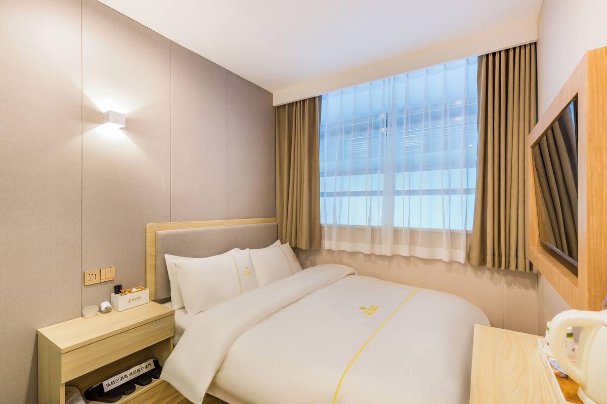 A hotel room with a neatly made bed, a bedside table with a lamp, a small shelf with toiletries, and a window with curtains.