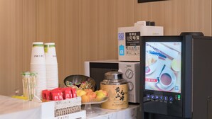 Zona de buffet con estación de bebidas de autoservicio, microondas y diversas opciones de comida, incluyendo frutas y aperitivos envasados.
