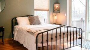 Une chambre à coucher comprenant un lit, une table de chevet, une fenêtre avec des stores, un luminaire fixé au mur et une vue sur l’extérieur.