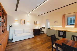 Um espaço compacto para viver, com sofá, mesa de jantar, piso de madeira, área de cozinha e estante de livros.