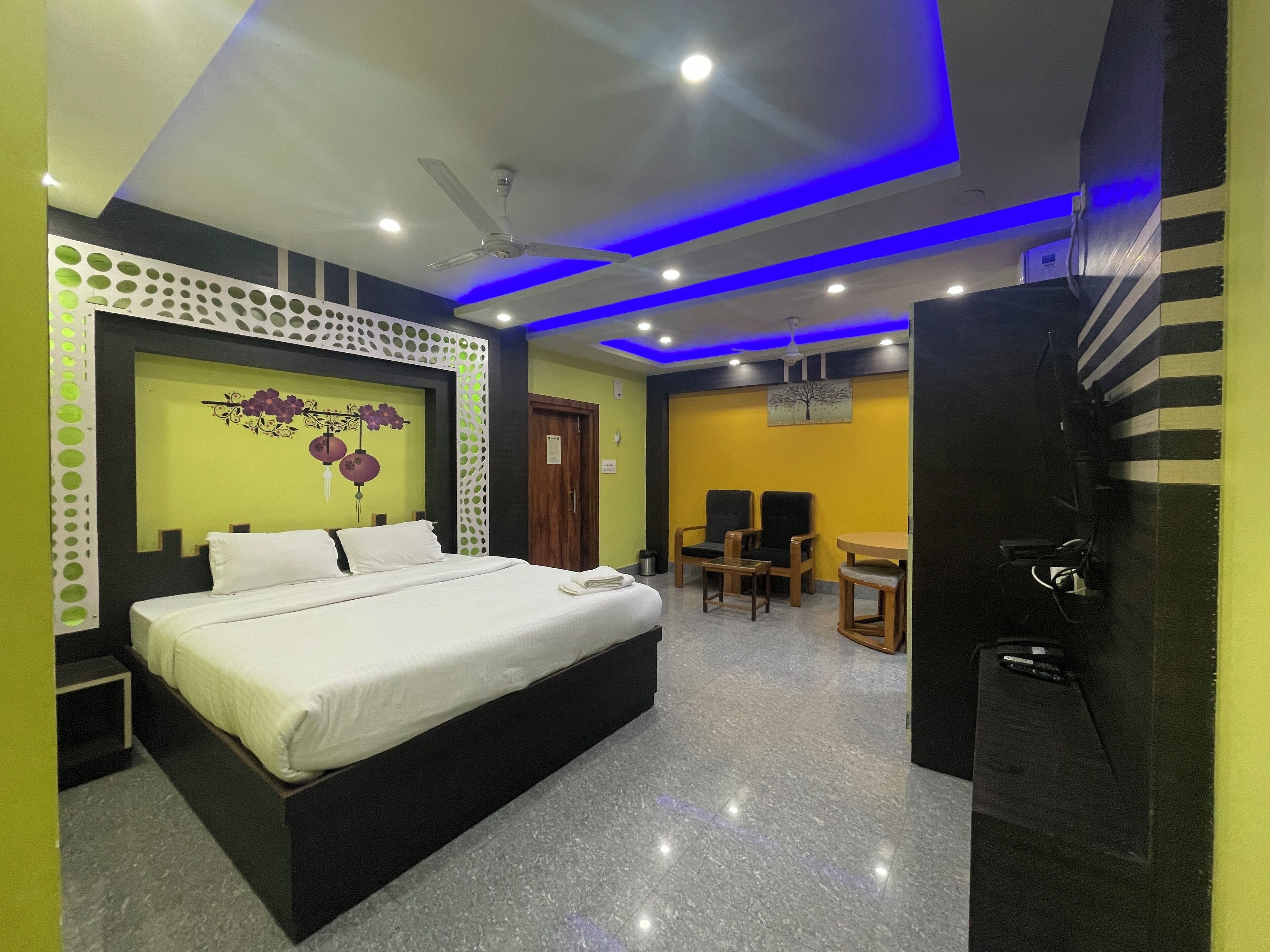 A hotel room with a bed, a desk, two chairs, a ceiling fan, and a TV.