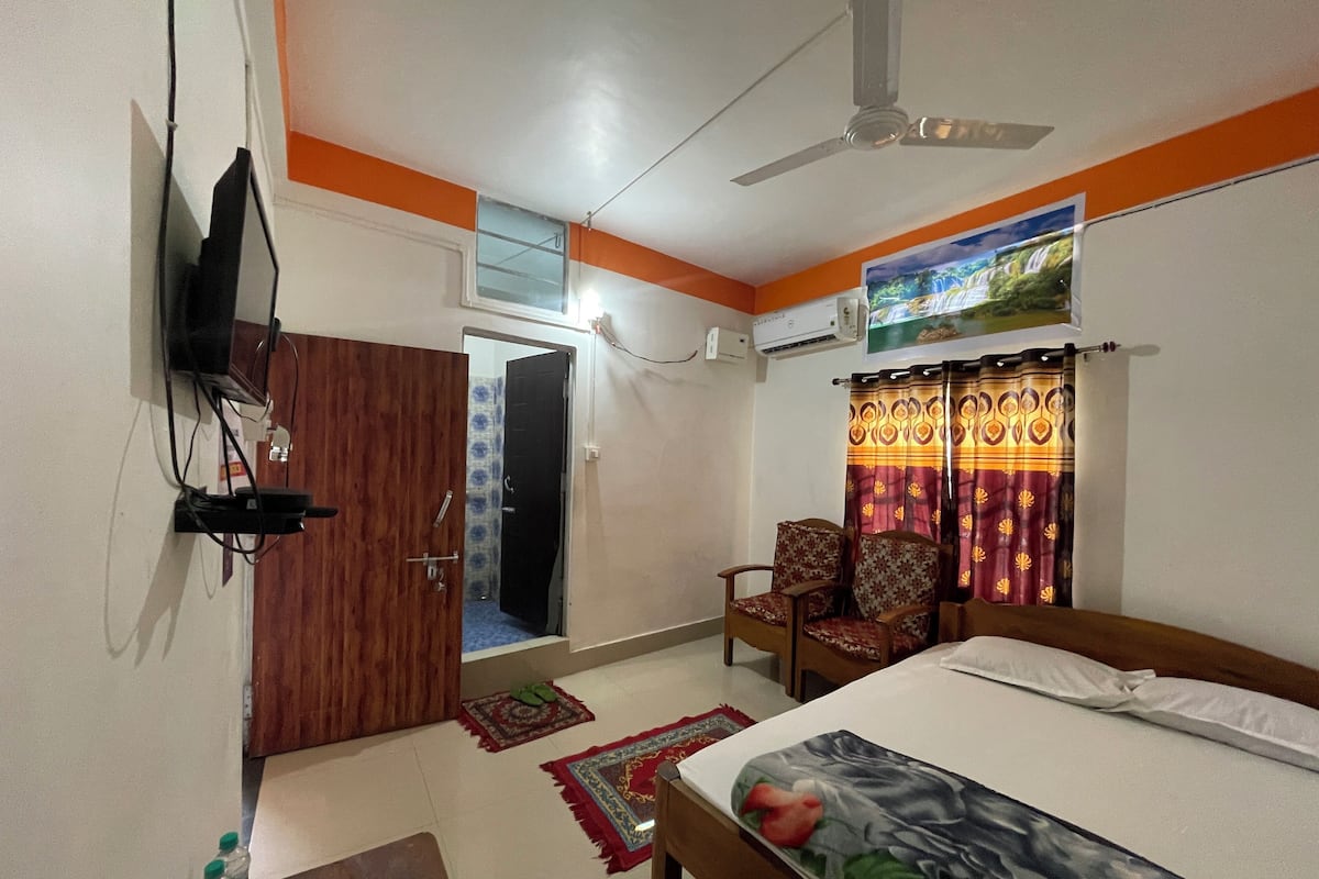 A hotel room with a bed, a ceiling fan, a TV mounted on the wall, a window with curtains, and a door.