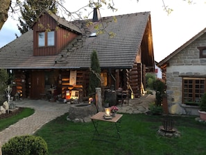 Une cabane rustique en rondins de bois, avec une extension en pierre, un espace extérieur aménagé et couvert, et un foyer.