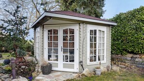 A small, white wooden structure with multiple windows, a stone pathway, and surrounding greenery.