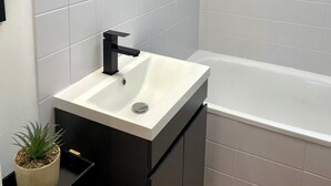 Ein modernes Badezimmer mit einem weißen Waschbecken, einem schwarzen Wasserhahn und einem Spiegel darüber. Auf der Ablage steht ein Topfpflanze und im Hintergrund ist eine Badewanne zu sehen.