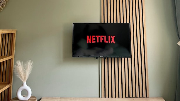 A modern living room with a mounted TV displaying "NETFLIX" on the wall, a wooden slat panel, a small round table with a vase, and a bed with a light-colored bedspread.