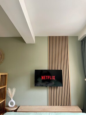 A modern living room with a mounted TV displaying "NETFLIX" on the wall, a wooden slat panel, a small round table with a vase, and a bed with a light-colored bedspread.