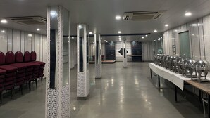 Un salón espacioso con filas de sillas rojas en un lado y una larga mesa con cubertería en el otro.