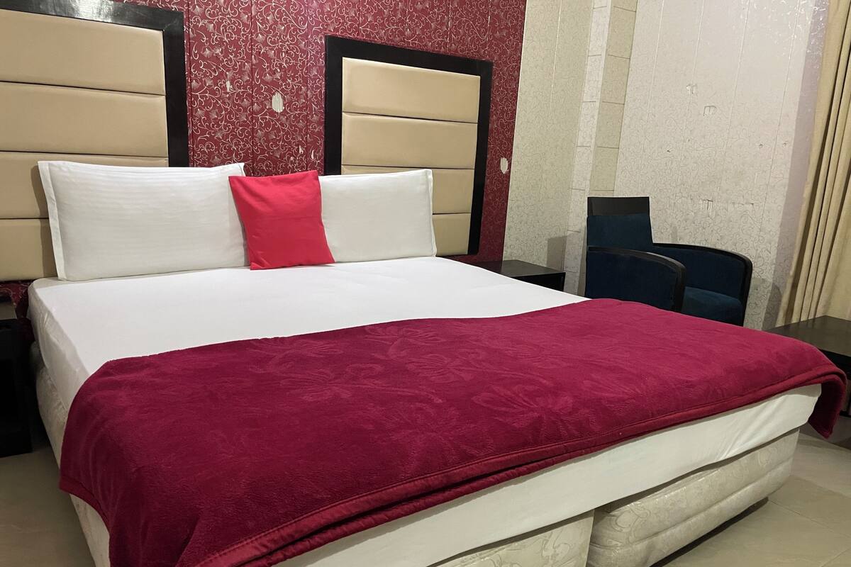 Un lit avec une couverture rouge, des oreillers blancs et un coussin décoratif rouge. Il y a une table de chevet avec une lampe, une chaise et un bureau avec un ordinateur. La pièce est décorée de papier peint à motifs et possède une fenêtre avec des rideaux.