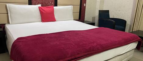 A bed with a red blanket, white pillows, and a red accent pillow. There is a nightstand with a lamp, a chair, and a desk with a computer. The room has patterned wallpaper and a window with curtains.