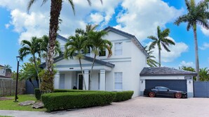 A two-story house with a garage, surrounded by palm trees and a well-maintained lawn. - Miami 5BR Pool Home ● BBQ ● Pool ● Coffee Bar ● Kid-Friendly ● Whirlpool Tub (Miami)