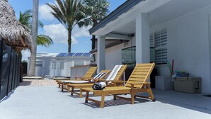 A beachfront property with wooden lounge chairs, a thatched-roof structure, and a modern building with solar panels. - Miami 5BR Pool Home ● BBQ ● Pool ● Coffee Bar ● Kid-Friendly ● Whirlpool Tub (Miami)