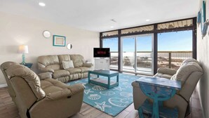 A living room with a beige sofa set, a coffee table, and a TV. There are large windows with a view of the beach.