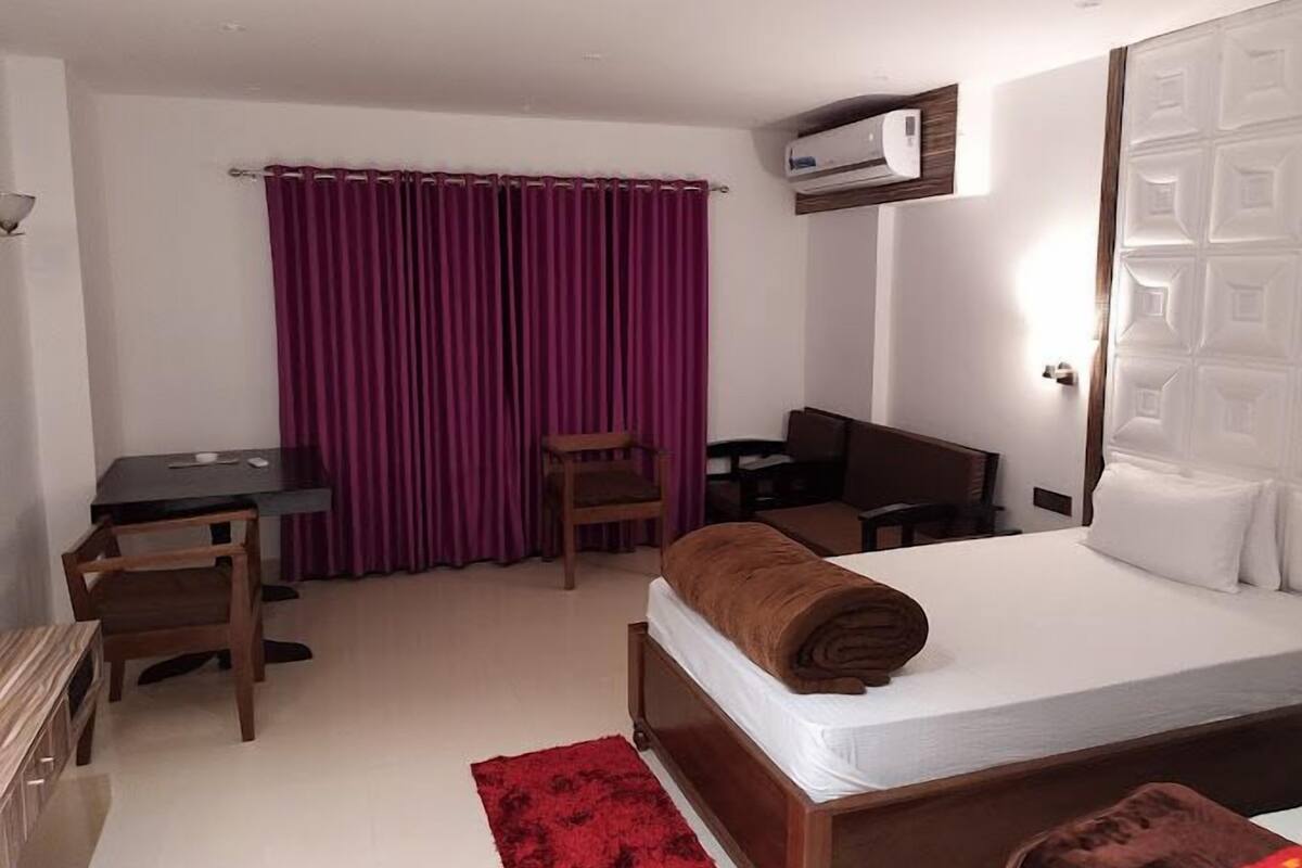 Une chambre d’hôtel avec un lit, un bureau, deux chaises, un tapis rouge et un appareil de climatisation.