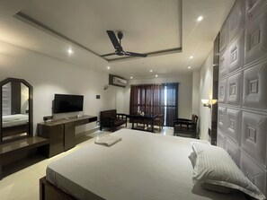 A hotel room with a bed, a desk, a chair, a TV, and a ceiling fan. - Hotel Living Hub (North Lakhimpur)