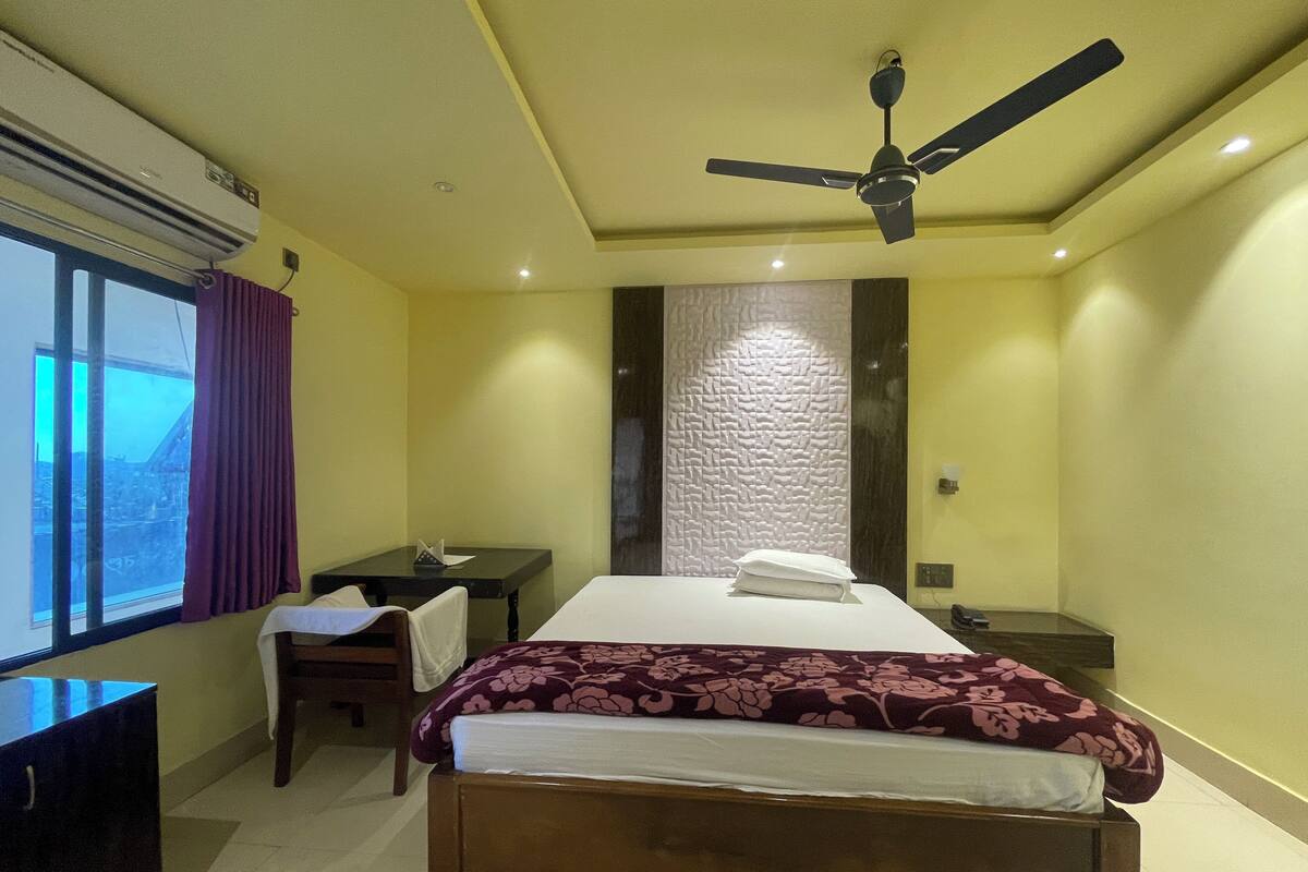 Une chambre d’hôtel comprenant un lit, un bureau, une chaise, une fenêtre avec des rideaux, un ventilateur de plafond et un panneau mural texturé.