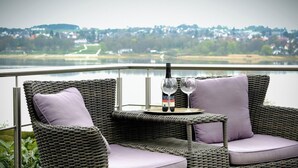 Des meubles en osier agrémentés de coussins, une bouteille de vin et un verre sur un balcon donnant sur un lac et une ville.