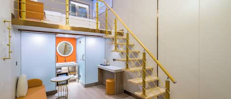 Una habitación de hotel moderna con cama tipo altiloja, una kitchenette pequeña, un espejo redondo, una escalera de madera y un área de descanso con una mesa redonda.