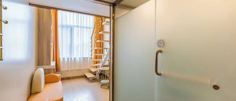 A modern hotel room with a glass partition, a sofa, a staircase, and a view of the outdoors.