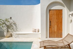 A poolside area with a wooden door, a lounge chair, a bench with cushions, and a potted plant.