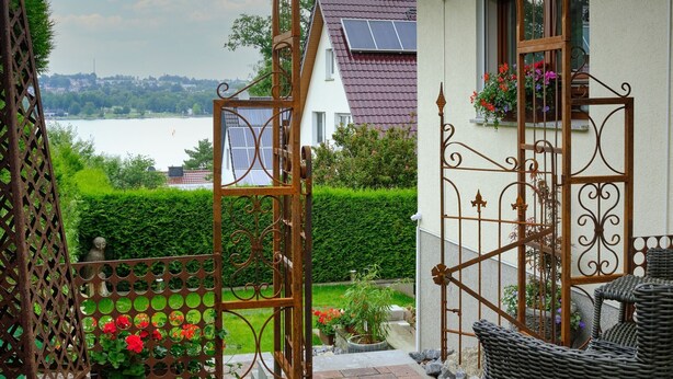 A gated entrance with a view of a lake, a solar panel on a house roof, and outdoor wicker furniture.