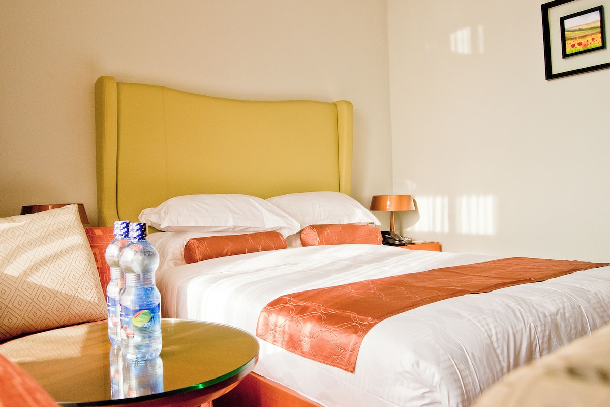 A hotel room with a bed, pillows, a bedside table with a water bottle, and a lamp.