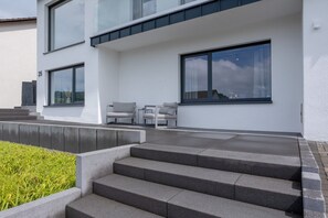 Un edificio moderno con una escalera que lleva a un balcón, mobiliario exterior y grandes ventanales.
