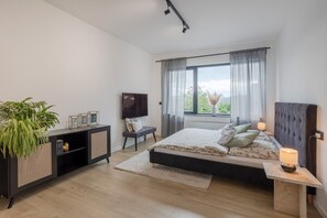 Ein modernes Schlafzimmer mit Bett, Nachttischen, einem Fernseher und einem großen Fenster mit Vorhängen.