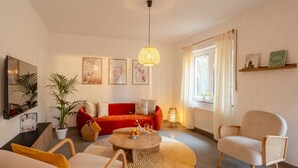 En moderne stue med en rød sofa, en beige lænestol og et sofabord. Der er to potteplanter, en vægmonteret reol med bøger og et stort vindue med gardiner.